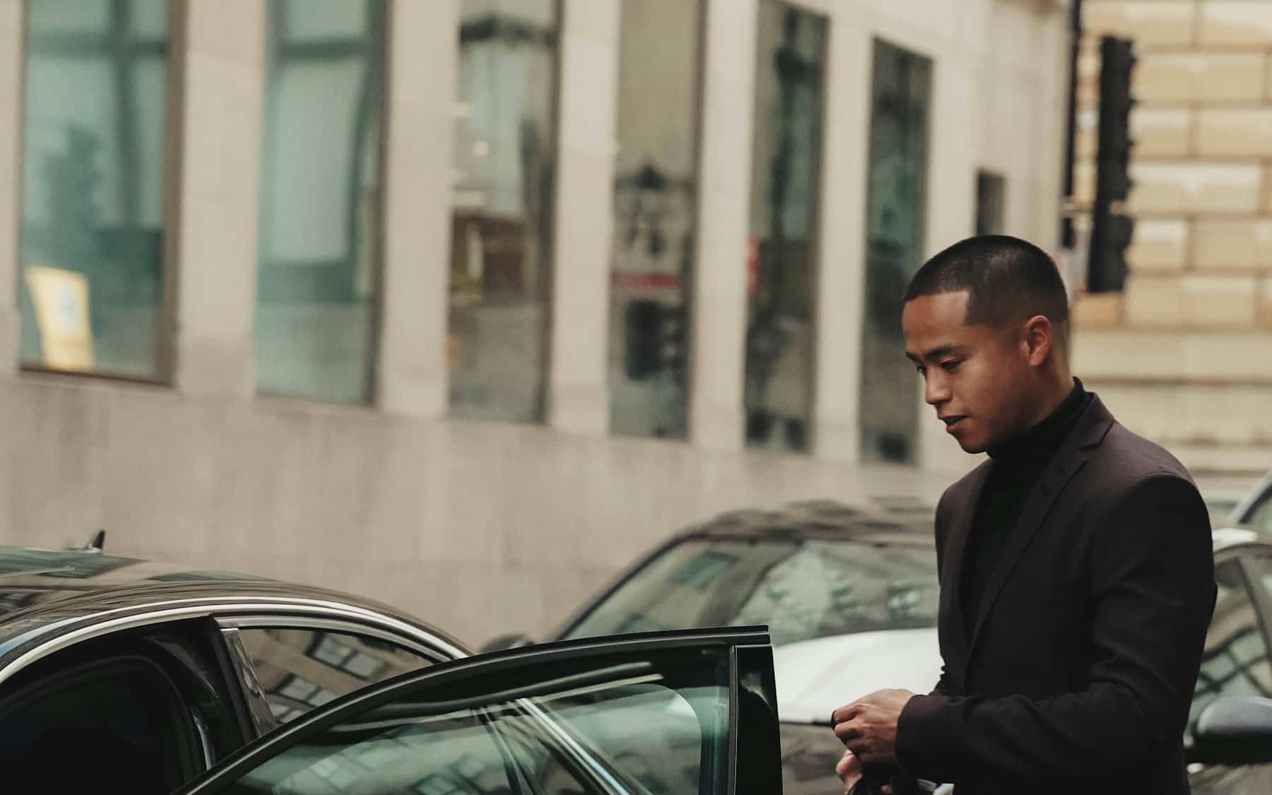A suited chauffeur holding open the door of a car for an arriving guest in London